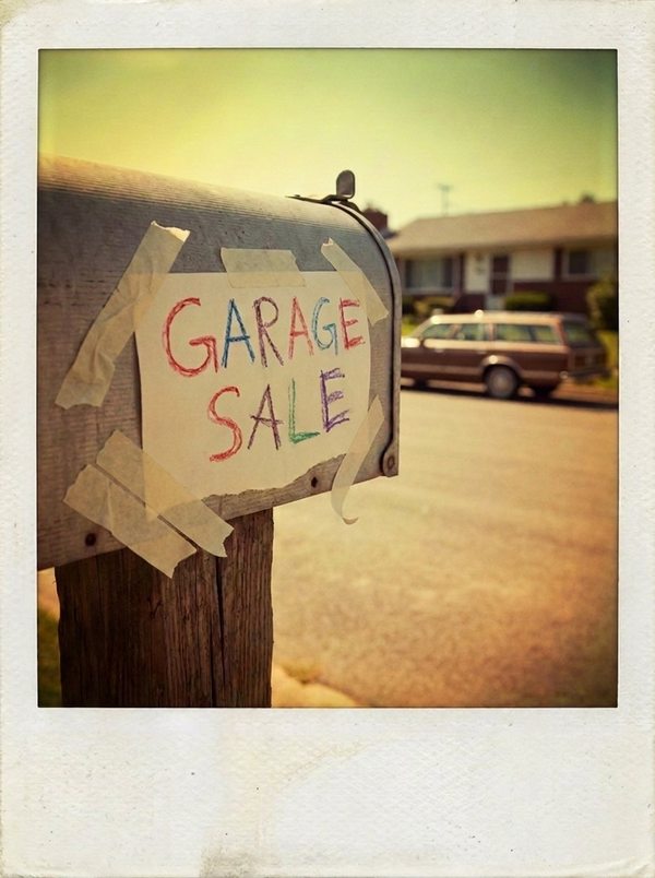 An AI recreation in the style of an old instant photo: a crayon sign reading GARAGE SALE taped to a mailbox at the end of a driveway, a station wagon across the street.